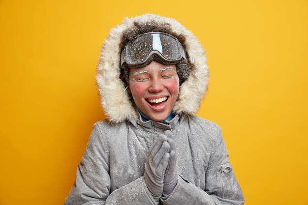 Beautiful happy woman hiker enjoys snowy winter and smiles broadly clasps hands has face covered with ice spends free time in mountains wears coat uses ski goggles.