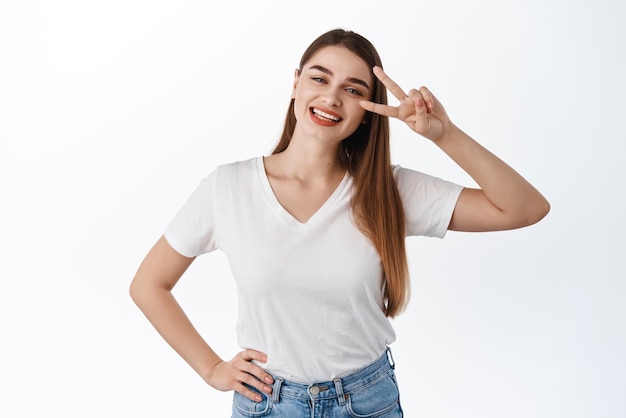 Beautiful happy woman shows peace vsign and smiles at camera joyful stay on bright side being optimistic standing in tshirt against white background