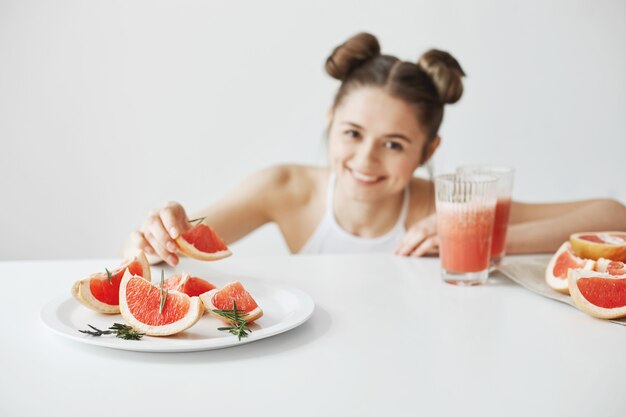 Beautiful happy woman smiling taking slice of grapefruit from plate sitting at table over white wall. Healthy fitness food.