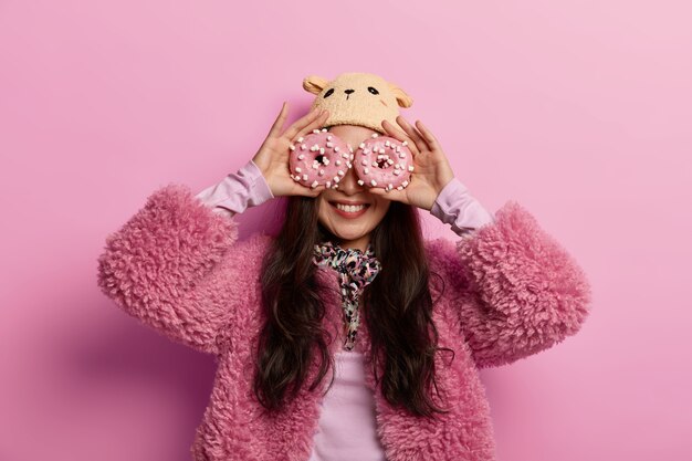 Free photo beautiful happy woman wears winter hat and coat, keeps glazed delicious doughnuts on eyes, likes eating confectionery