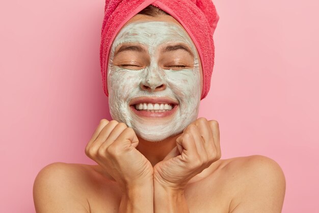 Beautiful happy woman with closed eyes, has clay mask on face, improves appearane, hydrates skin, smiles broadly, has white perfect teeth, feels being pampered like in spa, wears towel on wet hair