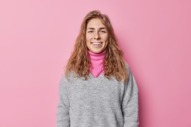 Beautiful happy woman with long hair smiles gladfully has clean natural skin ni make up feels happy wears casual jumper poses against pink background. Positive human face expressions and feelings