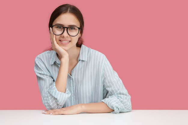 Beautiful happy young female employee wearing stylish blue shirt and spectacles enjoying working process,  with broad cheerful smile. People, lifestyle and occupation concept