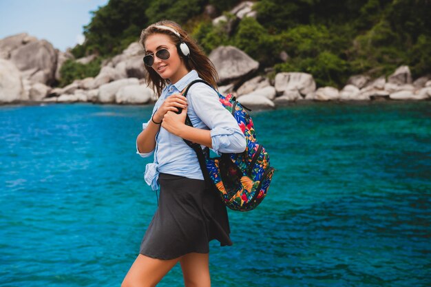 Beautiful hipster woman traveling around world with backpack, smiling, happy, positive, listening music in headphones, blue tropical ocean background, sunglasses, sexy, summer vacation,