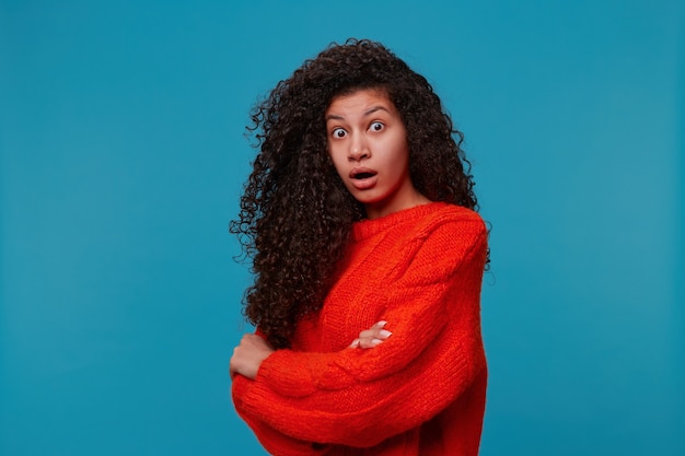 Beautiful hispanic latino woman stands half turn with arms crossed looks shocked amazed, with long dark curly wavy hair in red knitted sweater isolated over blue studio wall