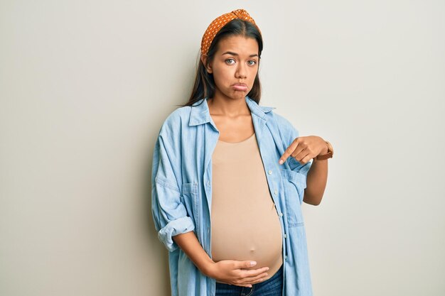 Beautiful hispanic woman expecting a baby, touching pregnant belly pointing down looking sad and upset, indicating direction with fingers, unhappy and depressed.