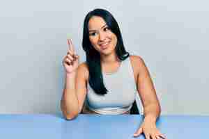 Бесплатное фото Красивая латиноамериканка с пирсингом в носу сидит на столе, показывая и указывая пальцем номер один, улыбаясь уверенно и счастливо.
