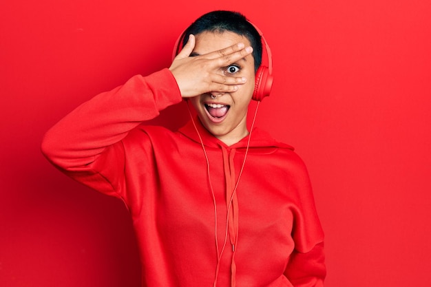 Free photo beautiful hispanic woman with short hair listening to music using headphones peeking in shock covering face and eyes with hand, looking through fingers with embarrassed expression.