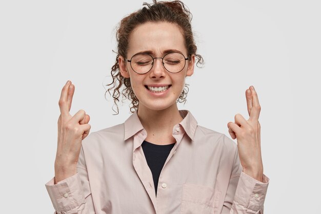 Beautiful hopeful dreamy young woman keeps fingers crossed, eyes closed, makes wish under shooting star, wears beige oversized shirt and spectacles, isolated over white blank wall