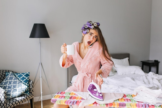 Free photo beautiful housewife in pink bathrobe at home. she ironing clothes, speaking on phone and holding a cup. she has curler on hair, smiling to camera