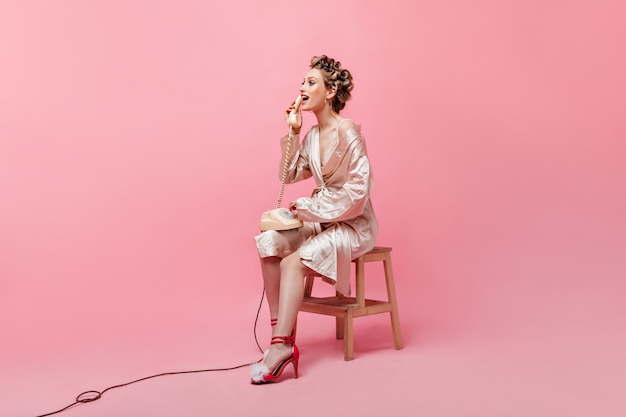 Красивая домохозяйка в розовом халате эмоционально разговаривает по телефону на изолированной стене