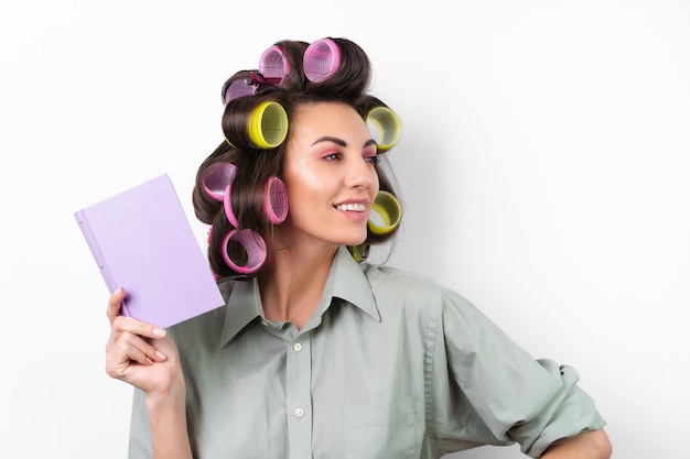 Foto gratuita bella casalinga giovane donna allegra con bigodini trucco luminoso con un libro in mano su uno sfondo bianco pensando a una ricetta per la cena alla ricerca di idee alimentari