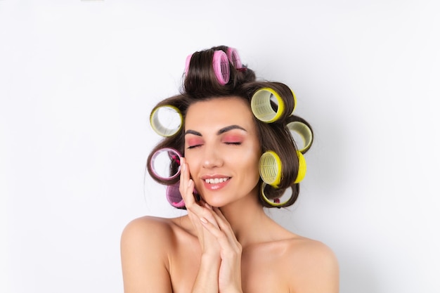 Beautiful housewife Young cheerful woman with hair curlers and bare shoulders getting ready for a date night Makes a hairstyle at home on a white background