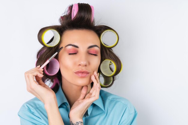 Beautiful housewife Young cheerful woman with hair curlers and eyebrow brush getting ready for a date night Makes a hairstyle at home on a white background