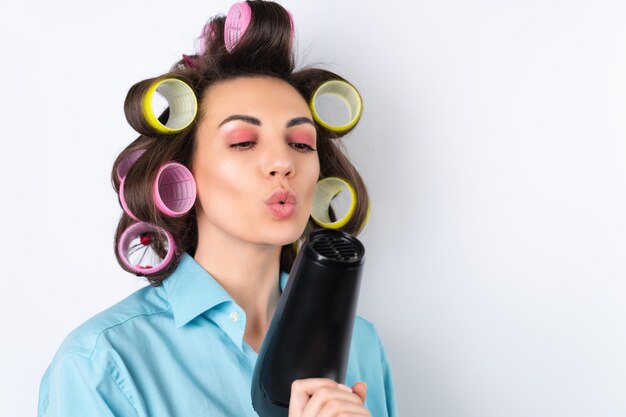 Beautiful housewife Young cheerful woman with hair curlers and hair dryer getting ready for a date night Makes a hairstyle at home on a white background