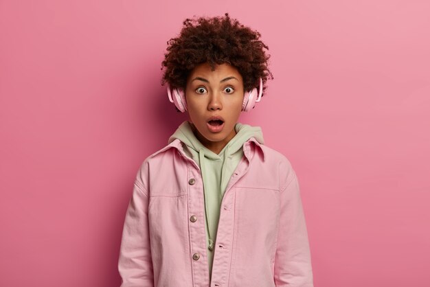 Beautiful impressed young woman has curly hair listens music with headphones, shocked by terrified relevation, wears pastel jacket, keeps mouth opened