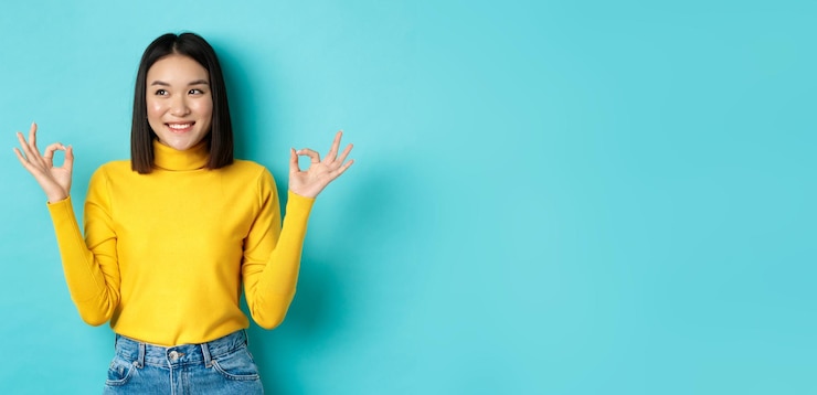 無料Photo 黄色のセーターを着た美しい日本人の女の子は、okサインを示し、左を向いて微笑み、ロゴとレックを示しています