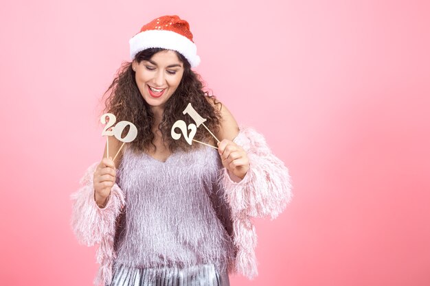 Beautiful joyful young brunette woman with curly hair in a Christmas cap on a pink background holding a wooden number for the new year concept