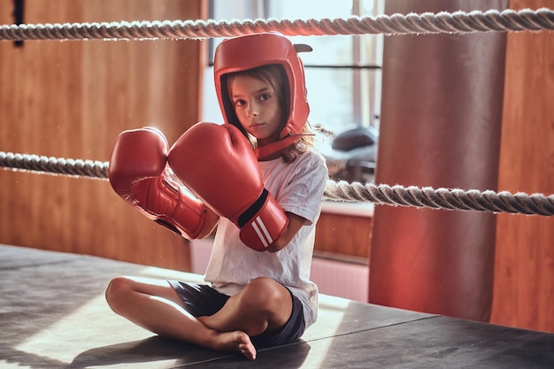 Beautiful kid girl is sitting on boxing ring wearing boxer uniform - gloves and helmet.
