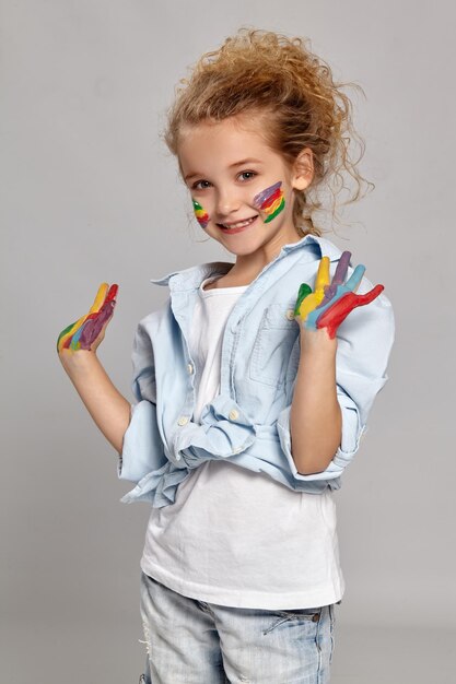 Beautiful kid having a brush in her chic curly blond hair, wearing in a blue shirt and white t-shirt. She is showing her painted arms and cheeks, smiling at the camera on a gray background.