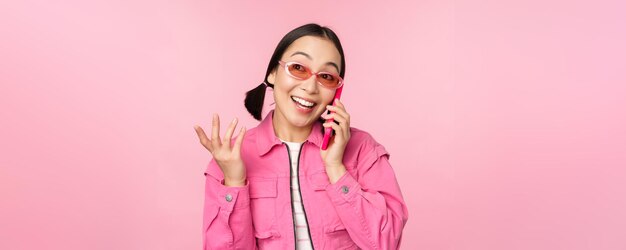 ピンクの背景の上に立っているスマートフォンで友人を呼び出すために携帯電話サービスを使用して幸せそうな顔で携帯電話で話しているサングラスの美しい韓国の女性モデル