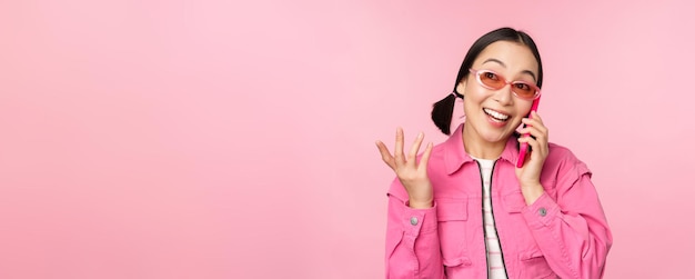 Free photo beautiful korean female model in sunglasses talking on mobile phone with happy face using cellular service to call friend on smartphone standing over pink background