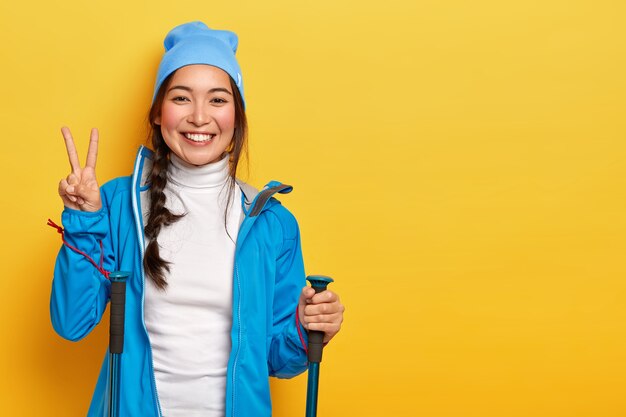 美しい韓国の女の子はハイキングを楽しんで、トレッキングスティックでポーズをとって、平和のジェスチャーをし、青い帽子とジャケットを着て、黄色の背景、空白のスペースで隔離