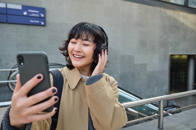 ヘッドフォンで美しい韓国の女の子は、携帯電話で通りに立っている彼女のスマートフォンでセルフィーを撮ります