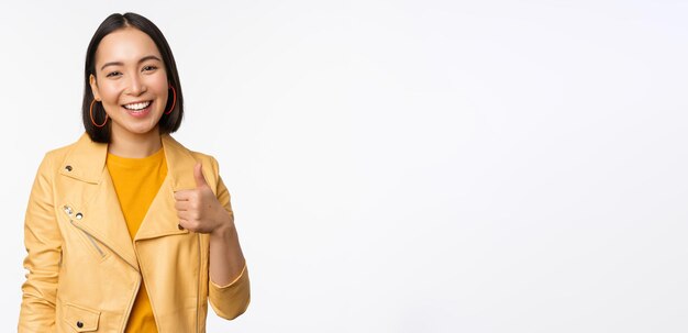 Beautiful korean girl smiling showing thumbs up like gesture recommending store or company standing satisfied against white background