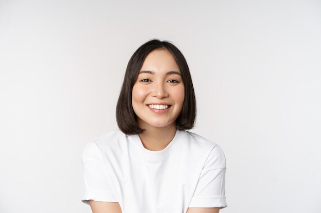 Beautiful korean girl smiling white teeth looking lovely at camera standing in white tshirt over studio background