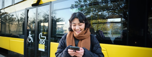 무료 사진 아름다운 한국 여자 학생이 버스 정류장에서 스마트폰을 보고 시간표를 확인하고 텍스트를 읽고 있습니다.