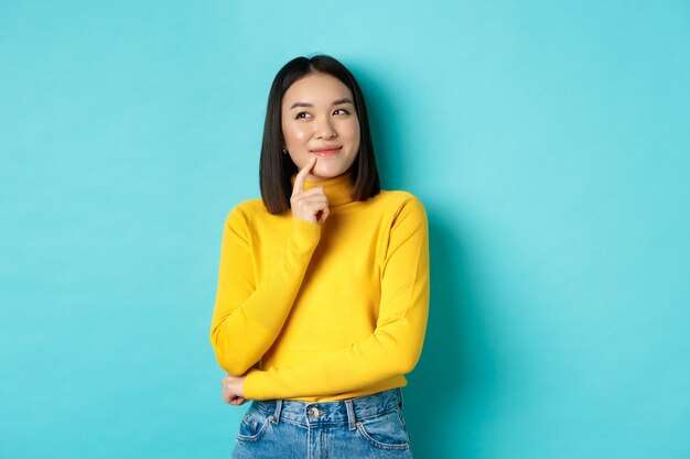 Beautiful korean girl thinking, imaging and smiling, looking at upper left corner and daydreaming, standing against blue background