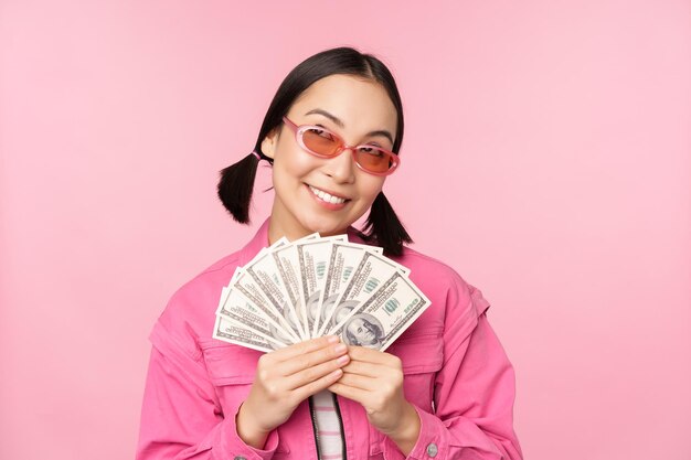 Beautiful korean woman in sunglasses showing dollars money cash smiling pleased concept of fast loans microcredit and payment standing over pink background