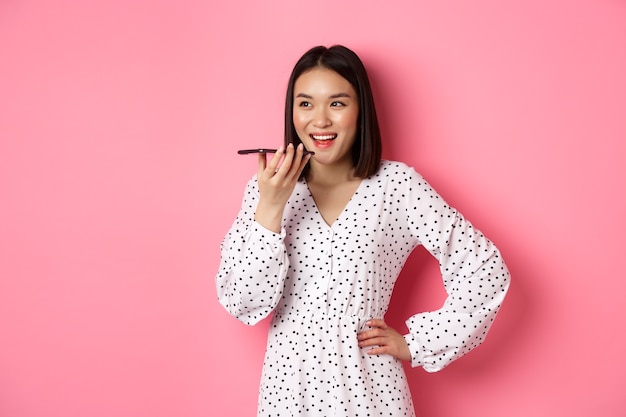 Beautiful korean woman talking on speakerphone, recording voice message and smiling happy, standing over pink background