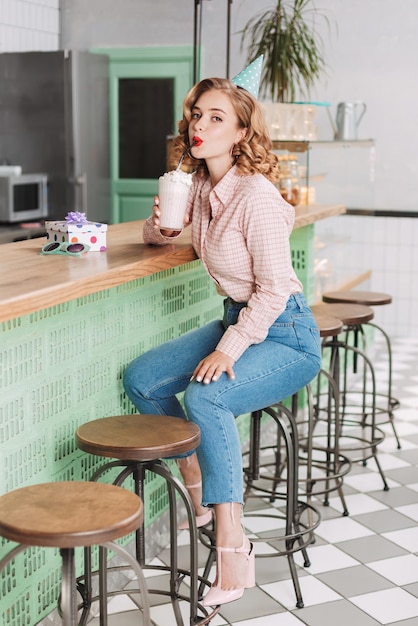 Foto gratuita bella signora con cappello di compleanno seduto al bancone del bar e bere milkshake mentre guarda sognante a porte chiuse nella caffetteria