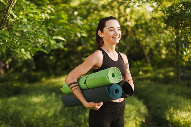 Beautiful lady in black sporty top and leggings standing with yoga mats in hands and happily looking aside in park