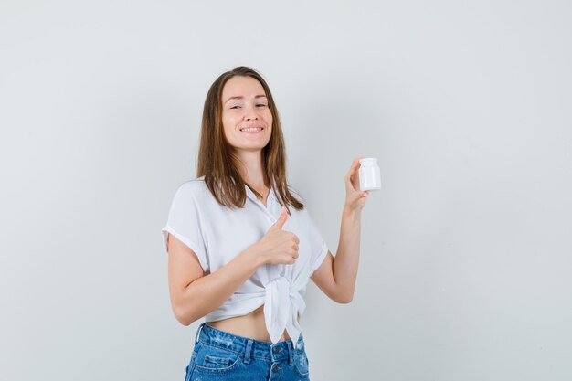 Beautiful lady holding bottle of pills while showing thumb up in white blouse,jeans and looking cheery , front view.