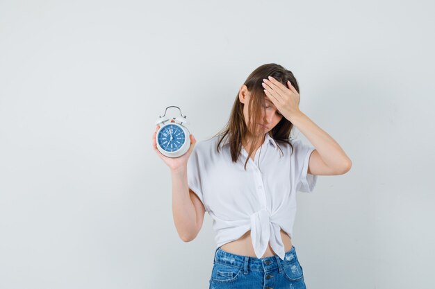 Beautiful lady holding clock while holding hand on head in white blouse and looking bored , front view.