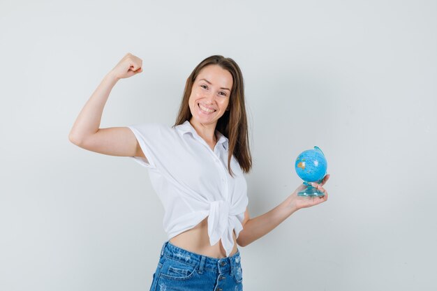 Beautiful lady holding globe while showing winner gesture in white blouse and looking cheery , front view.