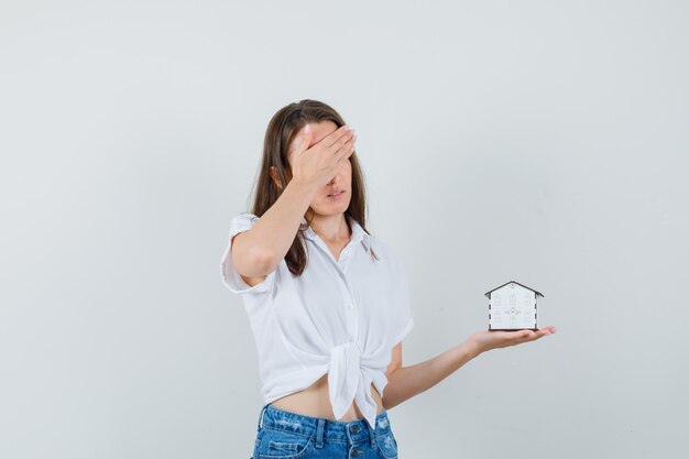 Beautiful lady holding model house while holding hand on her head in white blouse and looking stressful , front view.