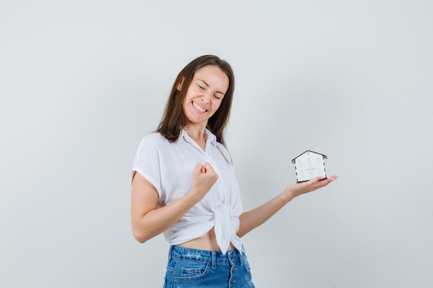 Foto gratuita bella signora che tiene la casa modello mentre mostra il gesto del vincitore in camicetta bianca e sembra felice, vista frontale.