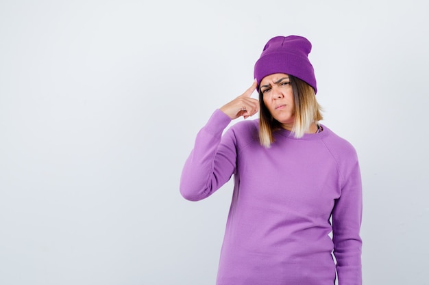 Beautiful lady keeping finger on head in sweater, beanie and looking puzzled , front view.