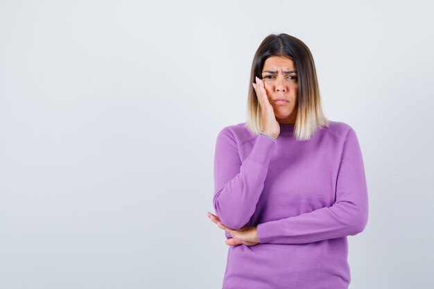 Beautiful lady keeping hand on cheek in sweater and looking dissatisfied , front view.