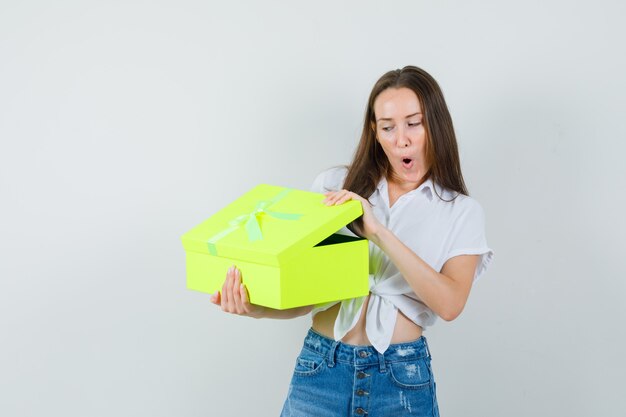 Beautiful lady opening gift box in white blouse,jeans and looking surprised , front view.