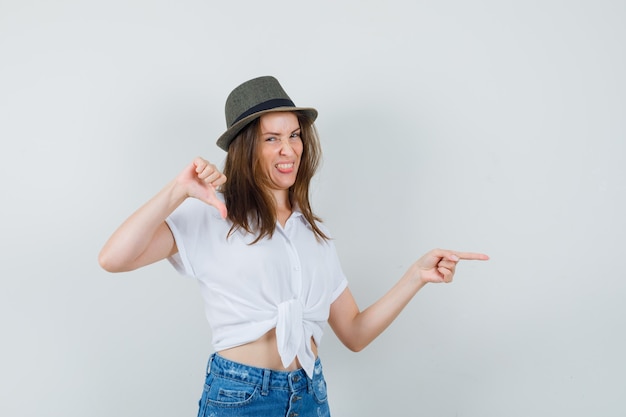 Beautiful lady pointing aside while showing thumb down in white blouse,hat , front view.