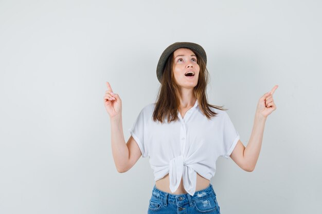 Beautiful lady pointing up in white blouse,hat and looking amazed , front view.