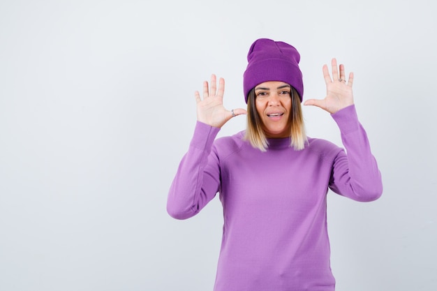 Beautiful lady raising hands in surrender gesture in sweater, beanie and looking cheerful. front view.