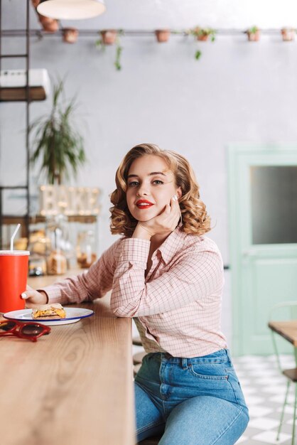 コカコーラグラスとバーカウンターに座って、カフェのカメラでドレミーに見ているシャツとジーンズの美しい女性