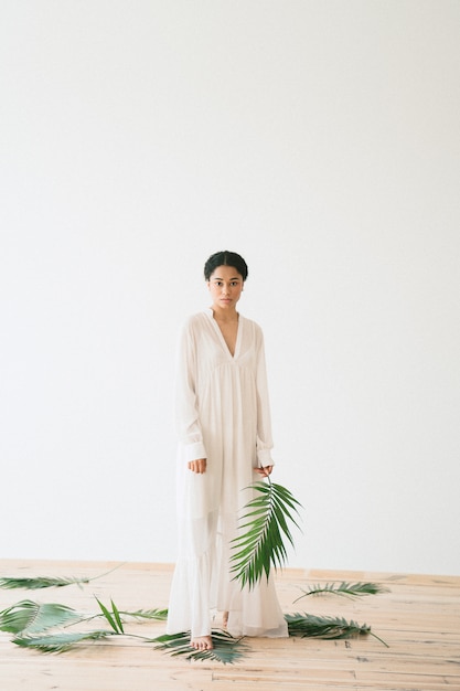 Beautiful lady standing looking and holding palm branch in long white dress in empty room . space for text