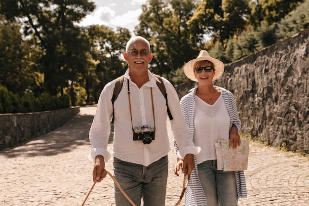 Beautiful lady in sunglasses, hat and striped blouse smiling and posing with man with mustache in white shirt and jeans with camera outdoor.